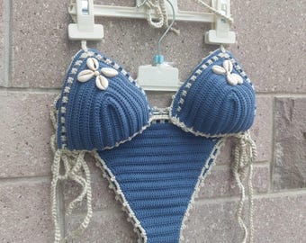May include: A handmade blue crochet bikini with seashell accents. The bikini top features seashell embellishments and a scalloped edge. The bottoms are a classic triangle cut. The bikini is displayed on a white hanger against a brick wall.