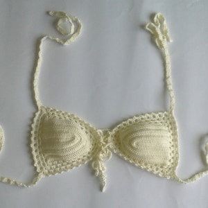 May include: A white crocheted bikini top with adjustable straps. The top has a lace trim and a tie in the front.