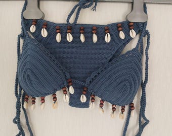 May include: A blue crochet bikini set. The top and bottom are adorned with small seashells and wooden beads. The bikini is hanging on a metal hanger against a white background. The crochet work is visible.