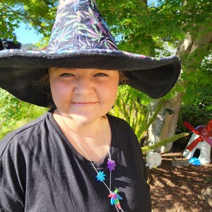 May include: A woman wearing a black witch hat with a colorful marijuana leaf print. She is also wearing a black shirt and a silver necklace with colorful marijuana leaf charms.