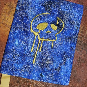 May include: A blue notebook with a gold skull design. The skull is dripping with gold paint. The notebook has a gold ribbon.