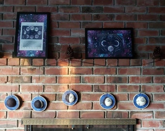 May include: A brick fireplace with a black metal screen and a wood-burning fire. Above the fireplace are six blue and white moon phase ornaments hanging from the ceiling. Two framed prints of moon phases are hanging on the brick wall above the ornaments.
