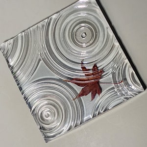 May include: A square, clear glass coaster with a raised design of concentric circles. A single, dried, red maple leaf is centered on the coaster.