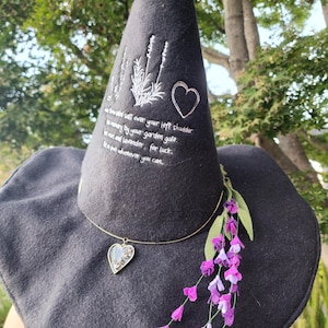 May include: Black witch hat with a wide brim and a tall pointed crown. The hat is decorated with white embroidery of lavender flowers and a heart. The text "Throw salted salt over your left shoulder when passing by your garden gate. Use rue and lavender for luck. Use it whenever you can." is embroidered on the hat. A gold chain necklace with a heart-shaped pendant is worn around the hat. The hat is adorned with a purple flower sprig.