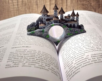 May include: A miniature castle bookend, featuring a detailed gray stone castle with brown turrets, sits atop an open book. The bookend spans the pages, creating a whimsical literary accent. The book is open to a page of printed text.