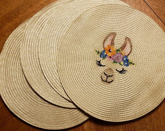 May include: Set of four round woven placemats. Each placemat features a beige woven design with a stitched llama face wearing a floral crown. The llama has brown ears, closed eyes, and a smiling mouth.