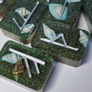 May include: Close-up of six square resin coasters with a silver metal design and seashells embedded in the resin. The coasters have a green and brown speckled background.
