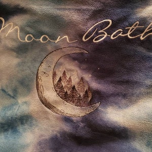 Può includere: Un tessuto tie-dye blu e bianco con le parole "Moon Bath" ricamate in bianco. Una falce di luna con una scena di foresta al suo interno è ricamata in marrone.