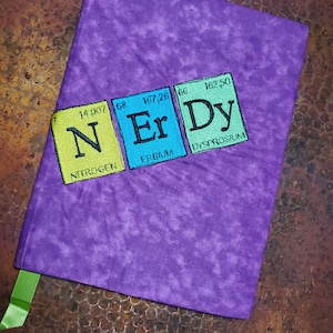May include: Purple fabric book cover with a green ribbon. The cover has an embroidered design of three colorful squares with the chemical symbols and names for nitrogen, erbium, and dysprosium.