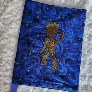 May include: A blue notebook cover with a galaxy print and an embroidered image of Groot from Guardians of the Galaxy. The cover has a blue ribbon attached.