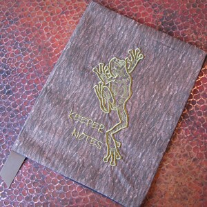 May include: A brown fabric notebook cover with a green embroidered frog design and the text "KEEPER NOTES".