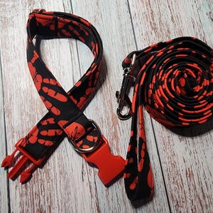 May include: A black and red dog collar and leash set. The collar and leash feature a repeating pattern of red handprints on a black background. The collar has a red plastic buckle and a metal D-ring. The leash is coiled.