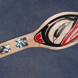 May include: A wooden paddle with a stylized design in black, red, and cream. The paddle features three inlaid abalone shell squares and a handle. The design depicts a bird or other creature.