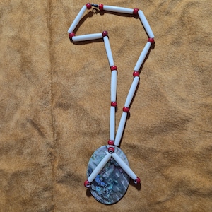 Può includere: Una collana fatta a mano con perline tubolari bianche e piccole perline rosse. La collana ha un ciondolo ovale con colori iridescenti. La collana è esposta su una superficie marrone.