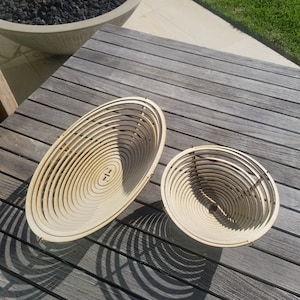 May include: Two wooden bowls, one larger than the other, are made of thin strips of wood that are stacked and curved to form a bowl shape. The bowls are light brown in color and have a natural wood finish.