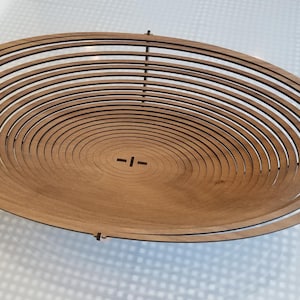 Può includere: Una ciotola di legno realizzata con sottili anelli concentrici di legno. La ciotola è di forma ovale e ha una finitura in legno naturale.