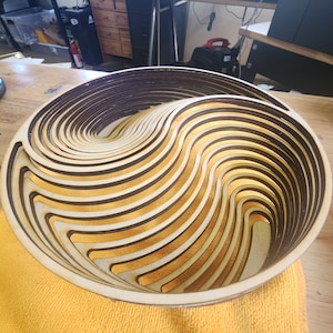 Könnte beinhalten: Eine Holzschale mit einem einzigartigen wellenförmigen Design. Die Schale besteht aus mehreren Holzschichten, die einen 3D-Effekt erzeugen. Die Schale ist hellbraun und hat eine glatte Oberfläche.