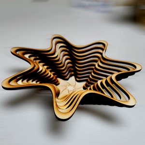 Puede incluir: Un cuenco de madera en forma de estrella con un diseño en capas. El cuenco está hecho de múltiples piezas finas de madera que se apilan una encima de la otra para crear un diseño único e intrincado.