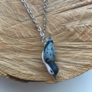 May include: A silver chain necklace featuring a bird pendant. The bird pendant is coloured white, black, and grey, with a detailed design. The necklace is displayed on a wooden surface.