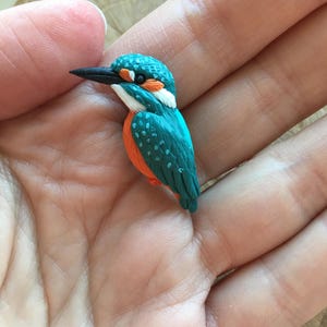 Könnte beinhalten: Eine handgefertigte Miniaturfigur eines Eisvogels. Der Vogel hat einen türkisfarbenen Körper mit weißen, orangefarbenen und schwarzen Akzenten. Die Figur wird in einer Hand gehalten, was die feinen Details und die leuchtenden Farben der Skulptur zeigt.