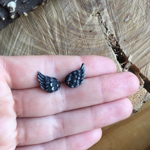 May include: A pair of small, black, detailed angel wing earrings. The wings have a textured, almost feathered look.