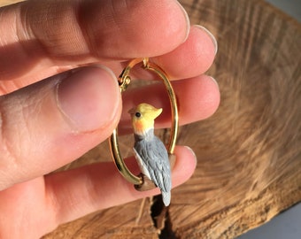 May include: Gold hoop earrings with a small, detailed ceramic cockatiel perched on a branch. The cockatiel is painted with yellow, white, and gray colors.