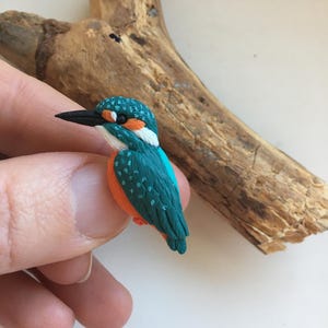 Könnte beinhalten: Eine handgefertigte Miniaturfigur eines Eisvogels. Der Vogel hat einen türkisfarbenen Körper mit orangefarbenen Akzenten, einen schwarzen Schnabel und eine weiße Brust. Die Figur wird in einer Hand gehalten, mit einem Stück Treibholz im Hintergrund.