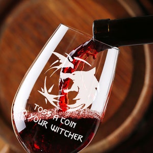 May include: A clear wine glass with a wolf graphic and the text "Toss a coin your Witcher" is being filled with red wine from a bottle.