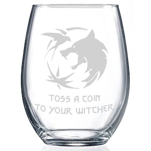 May include: A clear glass wine glass with an etched design of a wolf's head and the words "Toss a coin to your Witcher".