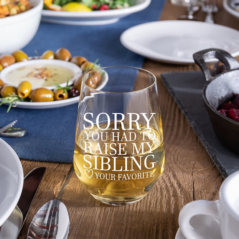 May include: A clear glass wine glass with the text "SORRY YOU HAD TO RAISE MY SIBLING YOUR FAVORITE" printed on it. The glass is filled with white wine and is sitting on a wooden table.