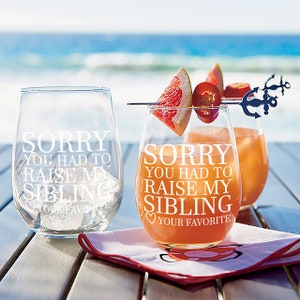 May include: Two clear stemless wine glasses with the text "Sorry you had to raise my sibling" etched on them. One glass has a grapefruit slice and a red pepper on a skewer. The glasses are on a wooden table with a red napkin.