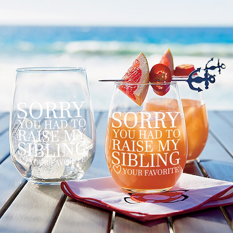 May include: Two clear stemless wine glasses with the text "Sorry you had to raise my sibling" etched on them. One glass has a grapefruit slice and a red pepper on a skewer. The glasses are on a wooden table with a red napkin.