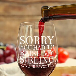 May include: A clear wine glass with the text "Sorry you had to raise my sibling" and a heart with the text "Your Favorite" printed on it. Red wine is being poured into the glass.