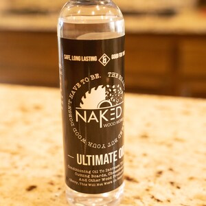 May include: A clear glass bottle with a black label that says "Naked Wood Works" and "Ultimate Oil". The label also has a saw blade graphic and the text "Doesn't have to be. The brand you don't have to hide."