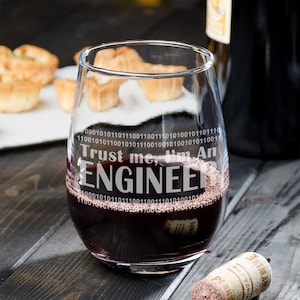May include: A stemless wine glass with the text "Trust me, I'm an ENGINEER" etched on the side. The glass is filled with red wine and is sitting on a wooden surface.