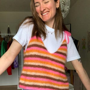 Può includere: Un top in maglia a righe multicolore, nei toni del rosa, arancione e marrone. Il top ha uno scollo a V e spalline annodate. È indossato sopra una t-shirt bianca. Lo stile è casual e fatto a mano.