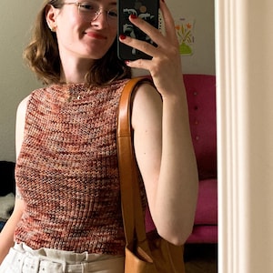 Könnte beinhalten: Eine Frau trägt ein braunes und oranges ärmelloses Oberteil mit einem weißen Gürtel und einer braunen Ledertasche. Sie steht vor einem Spiegel und hält ein schwarzes Smartphone.