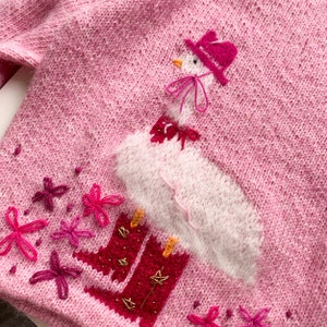 Könnte beinhalten: Rosa gestrickter Pullover mit einer weißen gestickten Gans, die einen rosa Hut und Stiefel trägt. Der Pullover ist mit rosa gestickten Blumen verziert.