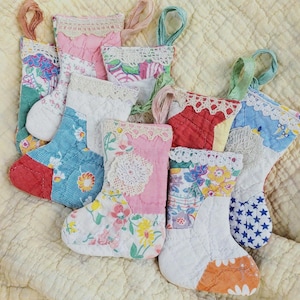 May include: A collection of seven small quilted stockings made from various patterned fabrics including floral, star, and solid prints. Each stocking features a decorative white lace trim at the top and a fabric loop for hanging purposes.