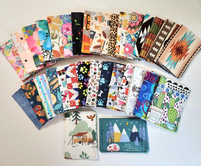 May include: A collection of rectangular fabric pouches featuring a wide variety of colorful patterns, including floral prints, animal motifs, geometric shapes, cactus designs, and outdoor scenes with campers and mountains, all arranged in a fan layout.