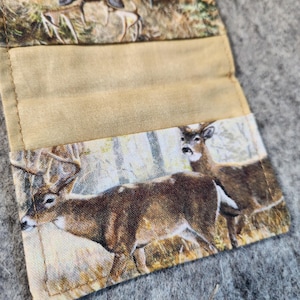 May include: A fabric card holder featuring a printed pattern of deer in a forest setting. The exterior displays a buck with antlers and a doe, while the interior has a solid tan fabric lining with stitched edges and a fold-over design.