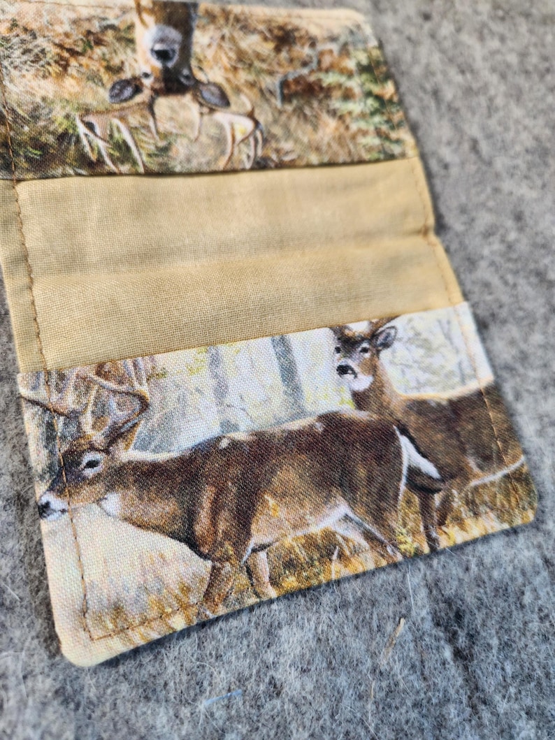 May include: A fabric card holder featuring a printed pattern of deer in a forest setting. The exterior displays a buck with antlers and a doe, while the interior has a solid tan fabric lining with stitched edges and a fold-over design.