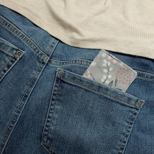 May include: A grey fabric wallet with a floral pattern featuring pink and white blossoms and green stems, tucked into the back pocket of blue denim jeans worn with a beige ribbed top. The wallet has a rectangular shape with rounded corners.