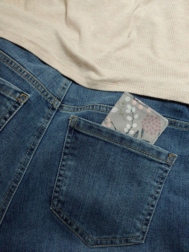 May include: A grey fabric wallet with a floral pattern featuring pink and white blossoms and green stems, tucked into the back pocket of blue denim jeans worn with a beige ribbed top. The wallet has a rectangular shape with rounded corners.