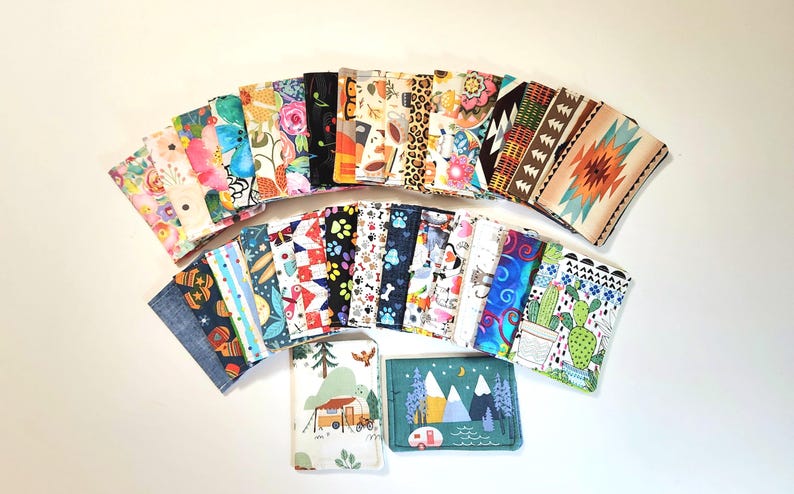 May include: A collection of rectangular fabric card holders arranged in a fan shape. The holders feature various colorful patterns including floral prints, geometric shapes, animal motifs, desert landscapes, and mountain scenes with campers.