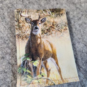 May include: A rectangular fabric patch featuring a printed image of a brown deer with antlers standing in a grassy field. The deer is positioned centrally, facing forward, with natural tones of tan, brown, and green visible in the detailed print.