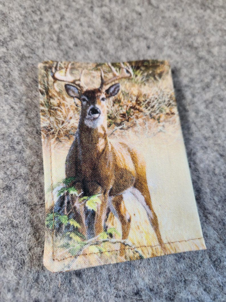 May include: A rectangular fabric patch featuring a printed image of a brown deer with antlers standing in a grassy field. The deer is positioned centrally, facing forward, with natural tones of tan, brown, and green visible in the detailed print.
