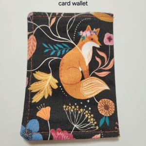 May include: A rectangular card wallet featuring a black fabric background with a printed pattern of an orange fox wearing a floral crown, surrounded by colorful flowers, leaves, and stems. The text Outside of card wallet is printed above.