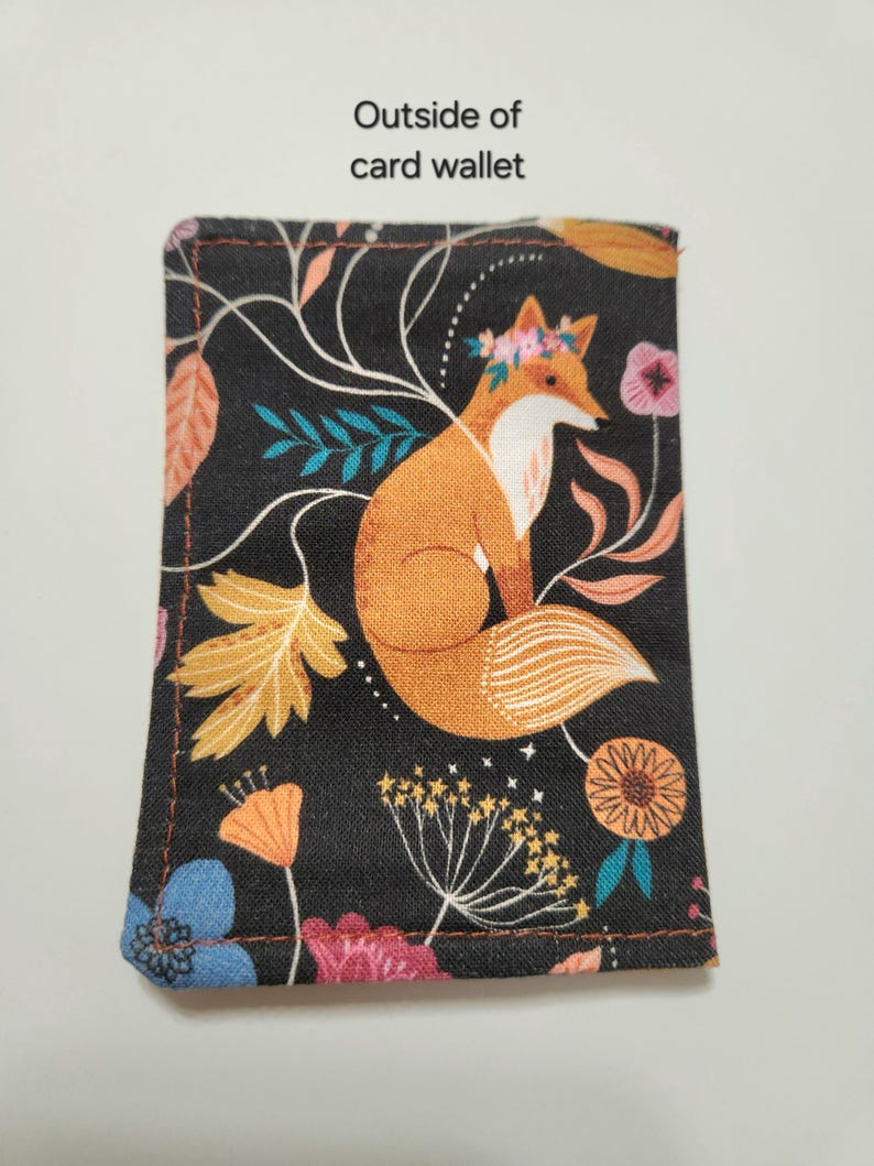 May include: A rectangular card wallet featuring a black fabric background with a printed pattern of an orange fox wearing a floral crown, surrounded by colorful flowers, leaves, and stems. The text Outside of card wallet is printed above.