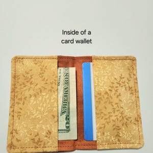May include: A fabric card wallet with a tan and brown leaf pattern. The interior features two pockets containing a twenty-dollar bill and a blue card. The text 'Inside of a card wallet' is printed above the open wallet on a plain background.